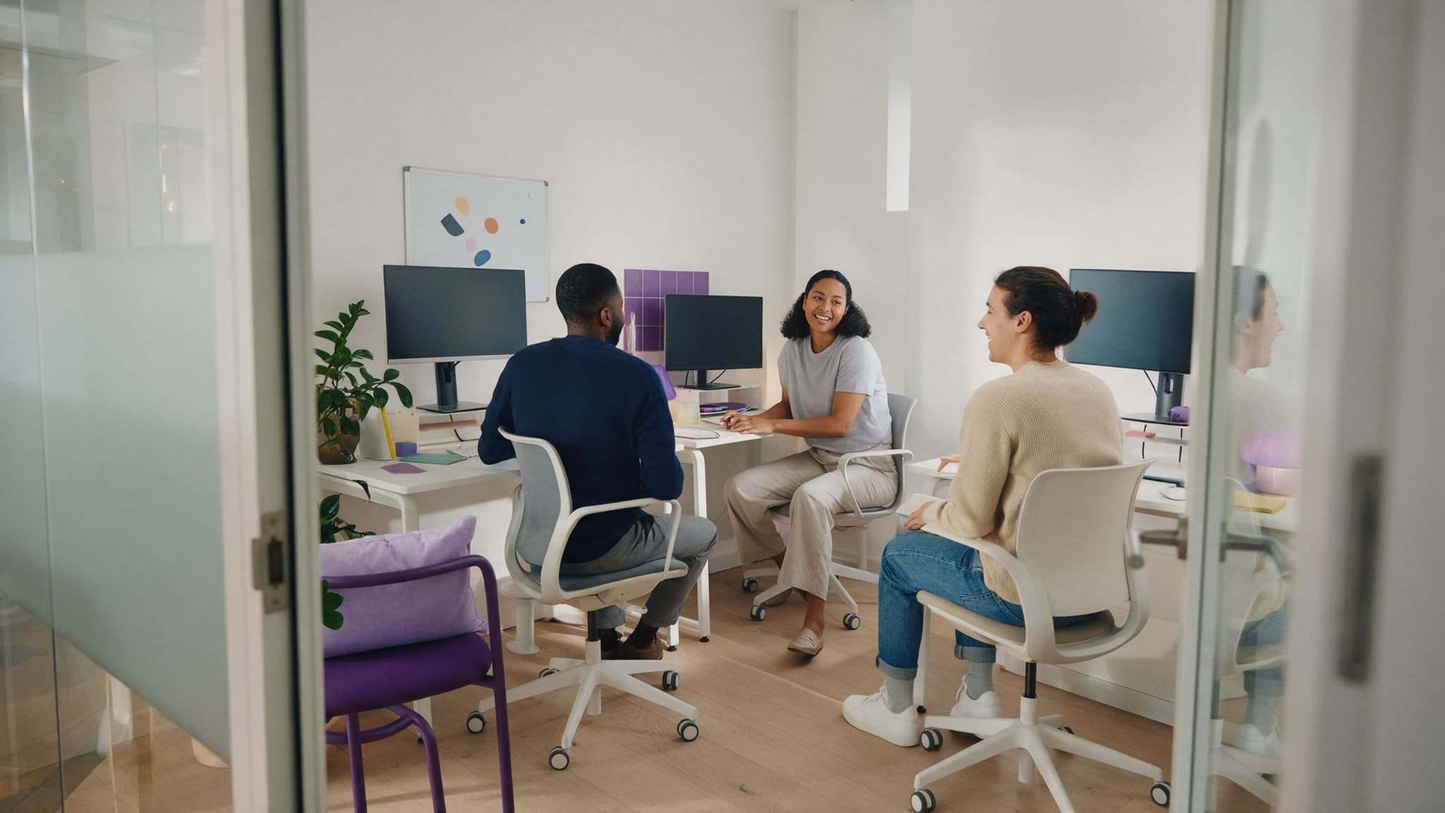 A team of three in its own small private office with glass walls and white desks; two work at their screens, one turns around to talk.
